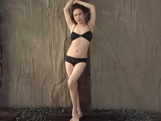 CockZilLaHotTs se tient adossée à un mur rugueux, les bras au-dessus de la tête, vêtue de lingerie noire avec un soutien-gorge et des culottes, peau pâle, cheveux bruns courts, avec une expression légèrement ennuyée. Elle est pieds nus sur du gravier..