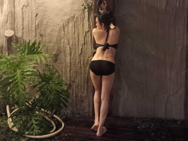 CockZilLaHotTs staat met de rug naar de camera, armen tegen een gestructureerde muur, draagt een zwart bikinie, toont slanke, bleke benen en een klein achterwerk. Een groene plant aan de linkerkant, houten vloer eronder..