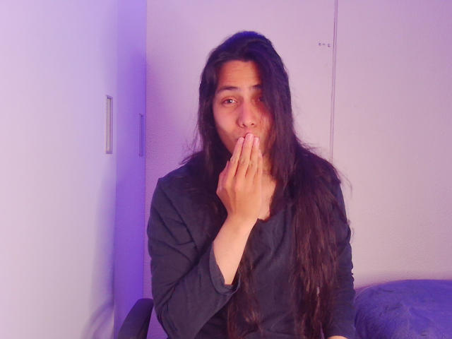 Una persona llamada Igorendon, con cabello largo y oscuro, cubriendo la nariz y la boca con la mano, vistiendo una camisa negra, con iluminación púrpura, sentada en un cojín azul, mirando a la cámara, con la mano parcialmente cubriendo el rostro..