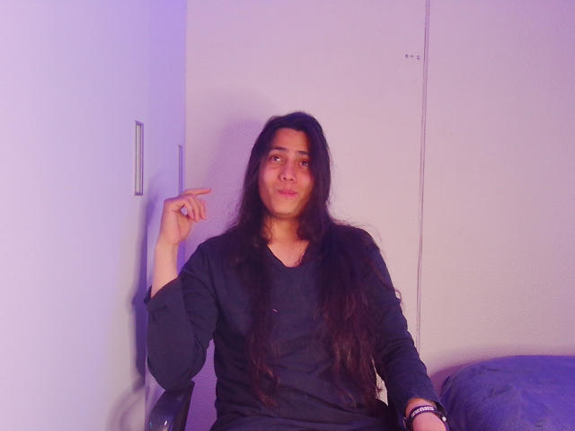 Igorendon with long dark hair, wearing a black shirt, sitting, pointing to the side with a slight smile, purple lighting, white wall, and a blue pillow.