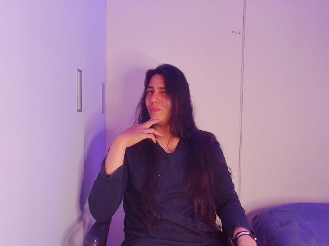 Igorendon, com longos cabelos escuros, vestindo uma camisa preta, está sentado com o dedo no queixo, parecendo um pouco entediado. Uma luz roxa ilumina suavemente seu rosto..