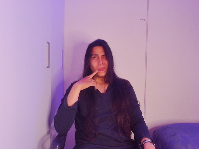 Een persoon genaamd Igorendon met lang donker haar, gekleed in een zwart shirt, zittend, met een vinger in de mond, in een paars-blauw licht, voor een witte muur, en op een grijze, beklede stoel, kijkend naar de camera..