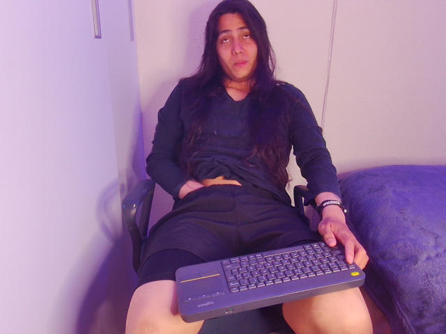 Igorendon, con cabello largo y negro, vistiendo una camisa negra de manga larga y pantalones cortos negros, está sentado en una silla sosteniendo un teclado, con las piernas separadas, y una almohada azul a su derecha..