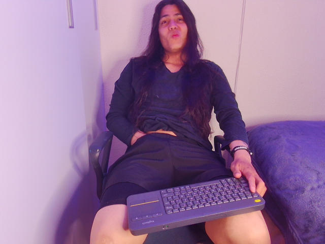 Igorendon mit langen schwarzen Haaren, einem schwarzen Hemd und einer schwarzen Shorts, sitzt auf einem Stuhl und hält eine Tastatur vor seinem Schritt. Er hat volle, rosafarbene Lippen. Im Hintergrund ist eine weiße Wand zu sehen..