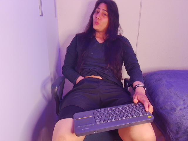 Igorendon, avec de longs cheveux noirs, un visage boudeur, portant une chemise noire et un short, tenant un clavier, assis les jambes écartées, dans une pièce éclairée en violet..
