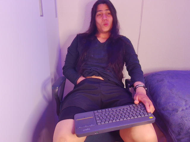 Igorendon, mit langem, schwarzem Haar, trägt ein schwarzes Langarmshirt und schwarze Shorts und sitzt mit einem Laptop im Schoß, wobei er/sie die Lippen zusammenpresst. Igoredons Hände ruhen auf den Oberschenkeln..