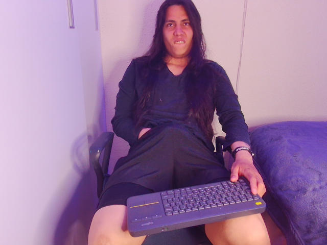 Igorendon with long black hair, wearing a black dress, sitting on a chair, holding a keyboard with hands in dress pockets, smiling, legs apart, blue blanket to the right.