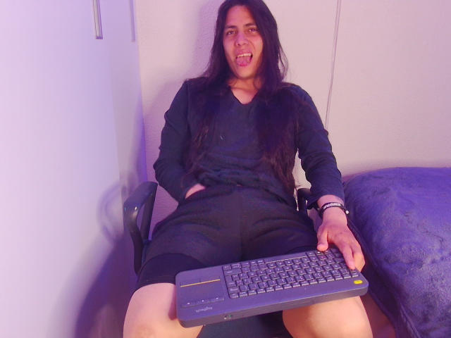 Igorendon mit langen, schwarzen Haaren, in einem schwarzen Kleid, sitzt auf einem Stuhl mit einer Tastatur vor sich, die Hände darauf, lächelt und hat die Zunge herausgestreckt. Eine blaue Decke liegt rechts daneben..