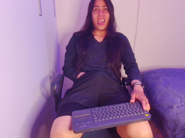 Igorendon mit langen, schwarzen Haaren, einem schwarzen Kleid und einer Tastatur, sitzt mit den Händen in den Taschen, der Zunge heraus und den Beinen gespreizt, und hält die Tastatur vor sich..