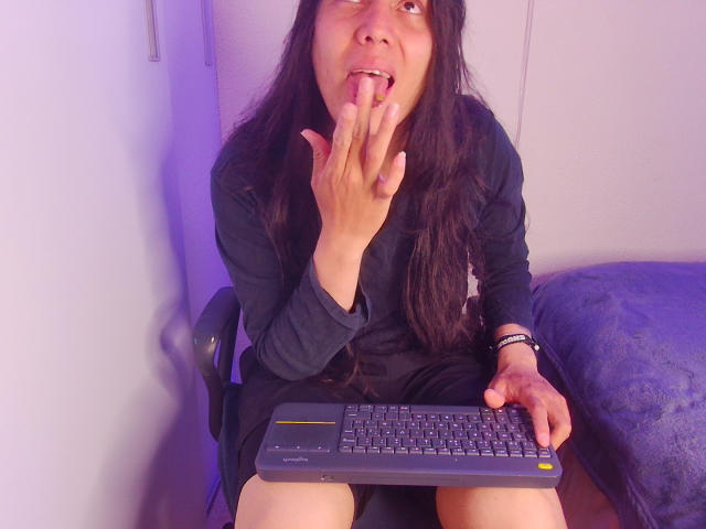 Igorendon, avec de longs cheveux noirs, portant une robe noire, assise sur une chaise, suçant son doigt, tenant un ordinateur portable, éclairage violet, couverture bleue, bracelet au poignet, regardant vers le haut..