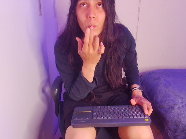 Igorendon, con el pelo largo y negro, vestida con un vestido negro, está sentada en una silla, haciendo pucheros y con el dedo en la boca, sosteniendo un teclado con la mano derecha, en una habitación iluminada con luz púrpura..