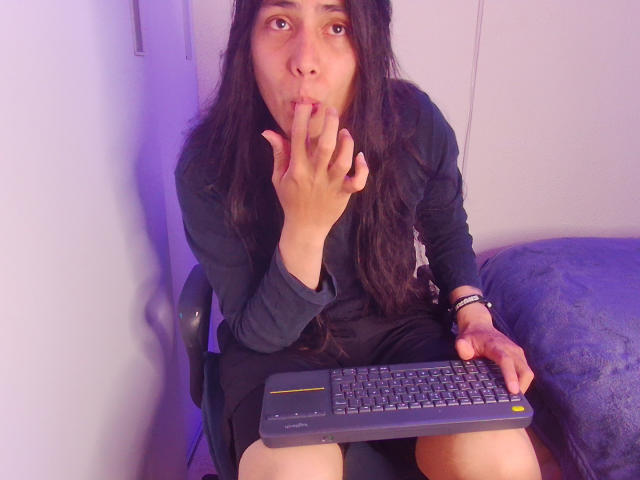 Igorendon with long dark hair, black shirt, and skirt, sucking their finger, holding a keyboard, right hand on keyboard, blue blanket to the right, purple light in background.