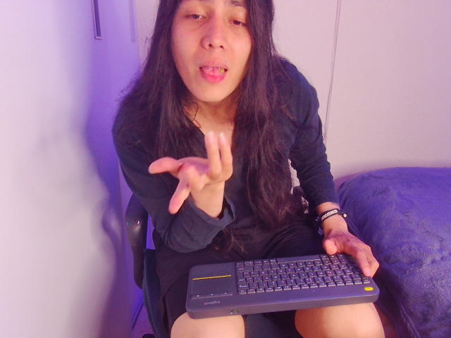 Igorendon mit langem, schwarzem Haar, der ein schwarzes Hemd trägt, sitzt auf einem Stuhl, hält eine Tastatur in der Hand, macht eine "pssst"-Geste mit den Fingern und hat den Mund leicht geöffnet..