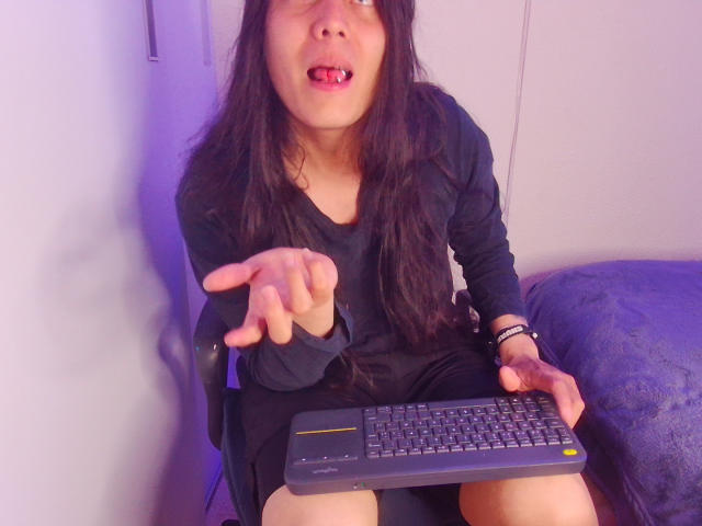 Igorendon, con el pelo largo y oscuro y la lengua afuera, señala un teclado con ambas manos, vistiendo una camiseta y pantalones cortos negros, y está sentado en una silla..