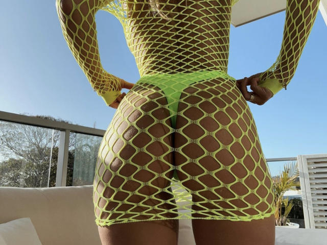 LilianaWax's ass in a neon yellow fishnet dress, revealing her green thong and bare butt. Her hands rest on her hips, highlighting her curvy, tan-skinned figure.