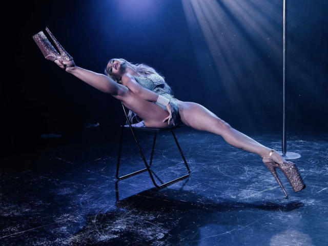 LilianaWax, mit weit gespreizten Beinen, hält glitzernde Schuhe, trägt ein glitzerndes Oberteil und Shorts, lehnt sich auf einem Stuhl zurück, unter blauem Scheinwerferlicht, mit einem Pfosten rechts..