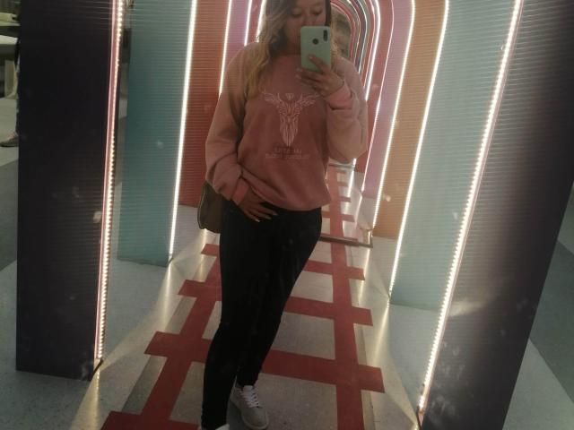 TamyParist draagt een roze sweater met een wit stierontwerp, zwarte broek en witte sneakers, en maakt een spiegelzelfportret met een groene telefoon..