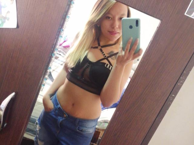 TamyParist, con cabello rubio, vestida con un top negro de tirantes que deja ver sus pechos y jeans azules, tomándose un selfie frente al espejo con su teléfono, con las manos en los bolsillos, sonriendo..