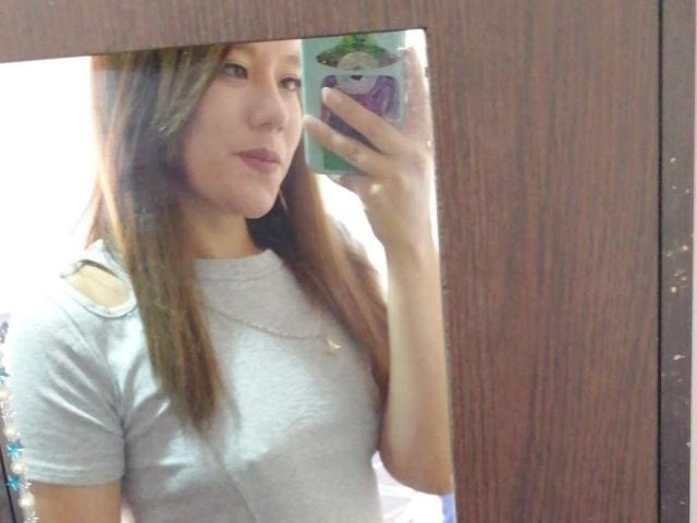 TamyParist, con i capelli castani, indossa una camicia grigia e tiene in mano un telefono con una custodia verde e viola. Il suo riflesso è visibile in uno specchio con cornice in legno, che mostra il suo viso e i suoi piccoli seni..