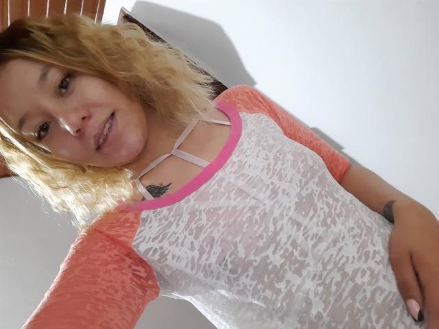 TamyParist, com cabelo loiro encaracolado, blusa de renda rosa e branca, seios pequenos visíveis, detalhes rosa, fundo branco, sorrindo, tatuagem no lado direito do pescoço, mão na cintura..