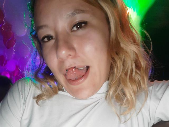 TamyParist with blonde wavy hair, white shirt, and silver tongue piercing, sticking out her tongue, has a slight smile, brown eyes, and a light skin tone.