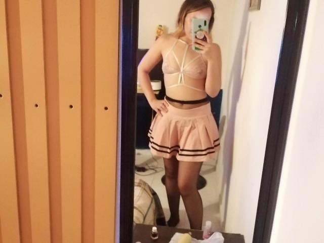 TamyParist indossa un reggiseno a fascia bianco trasparente con spalline sottili, che mette in mostra piccoli seni, e una minigonna rosa plissettata con strisce nere, mentre scatta un selfie allo specchio, tenendo in mano un telefono verde..