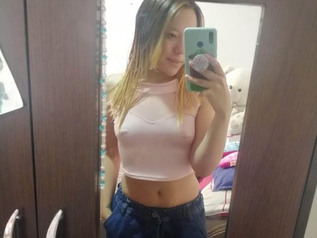 TamyParist, con i capelli biondi, indossa una maglia rosa trasparente che lascia intravedere i seni, e jeans blu, mentre scatta un selfie allo specchio, tenendo in mano un telefono verde con un pop socket..