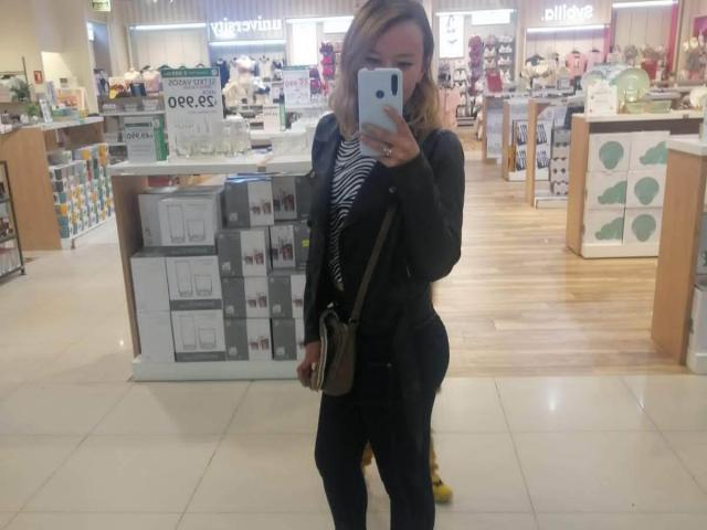TamyParist, con un seno di media grandezza, indossa un blazer nero, una camicia a righe e pantaloni neri, tiene un telefono in un negozio e si trova di fronte a un espositore con delle scatole..