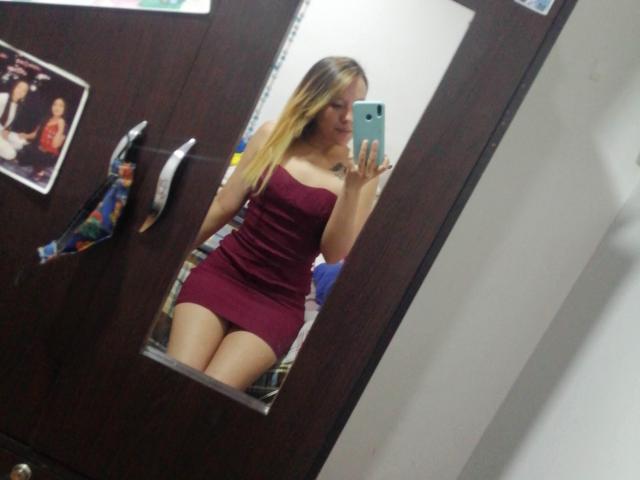 TamyParist indossa un abito viola senza spalline, aderente, che mette in mostra il suo sedere in un selfie allo specchio, tiene in mano un telefono blu, ha lunghi capelli biondi e si trova in una stanza con una porta di legno scuro..