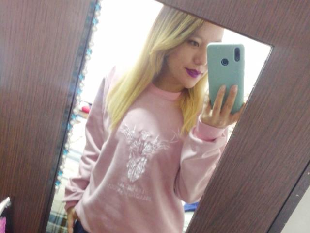 TamyParist, con i capelli biondi, un maglione rosa e rossetto viola, sta scattando un selfie allo specchio, tenendo in mano un telefono color acquamarina, mostrando leggermente il seno, in uno specchio con cornice di legno..