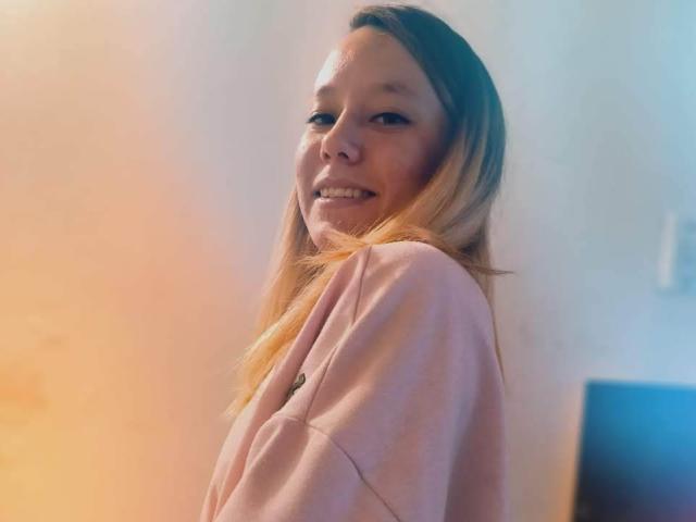 TamyParist, con cabello rubio, sonriendo, vestida con una camisa rosa, mirando por encima del hombro, mostrando su lindo trasero en la foto..