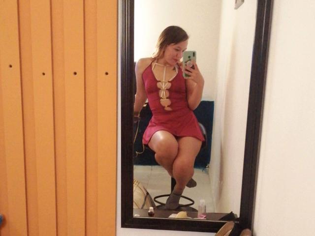 TamyParist com um vestido vermelho, exibindo o bumbum em uma selfie no espelho, usando um colar comprido, segurando um telefone, sentada em uma cadeira, com uma porta de madeira ao fundo..
