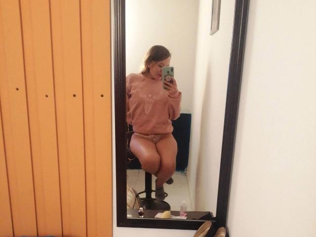 TamyParist, con cabello castaño medio, un suéter rosa y una tanga de encaje, mostrando su trasero y su vulva en un selfie frente al espejo, de pie sobre un taburete, sosteniendo un teléfono..