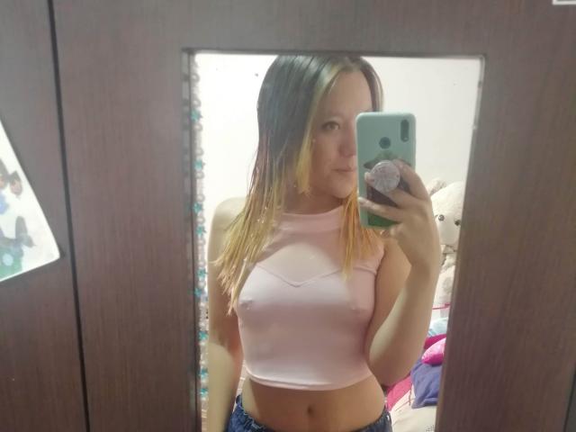 TamyParist, con un cabello de color rubio a castaño en degradado, vistiendo una camiseta rosa corta, mostrando sus pequeños senos, y sosteniendo un teléfono verde en un selfie frente al espejo..