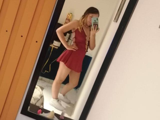 TamyParist, con cabello castaño largo, vestida con un vestido rojo, de pie frente a un espejo, sosteniendo un teléfono, con calcetines blancos, mostrando una figura delgada, linda, pequeña y con poco busto..