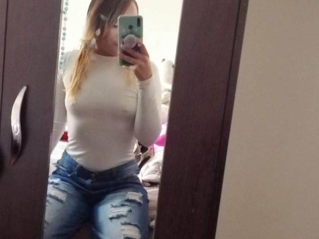 TamyParist with blonde hair in a white, tight, long-sleeve shirt, showing nipples, and ripped blue jeans, taking a mirror selfie with a green phone, in a bedroom.