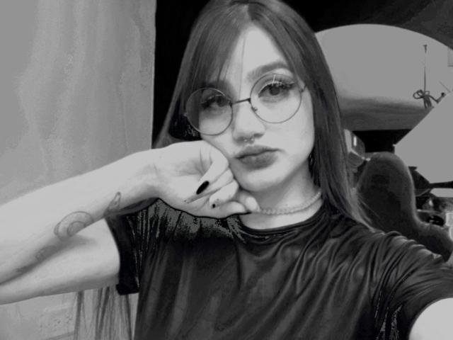 Gaby Durand mit langen Haaren, Brille, Nasenring und schwarzem Hemd, die ihr Kinn in die Hand legt, zeigt ein Tattoo am Handgelenk, blickt in die Kamera, Schwarz-Weiß-Foto, weiches Licht, subtile Mimik..