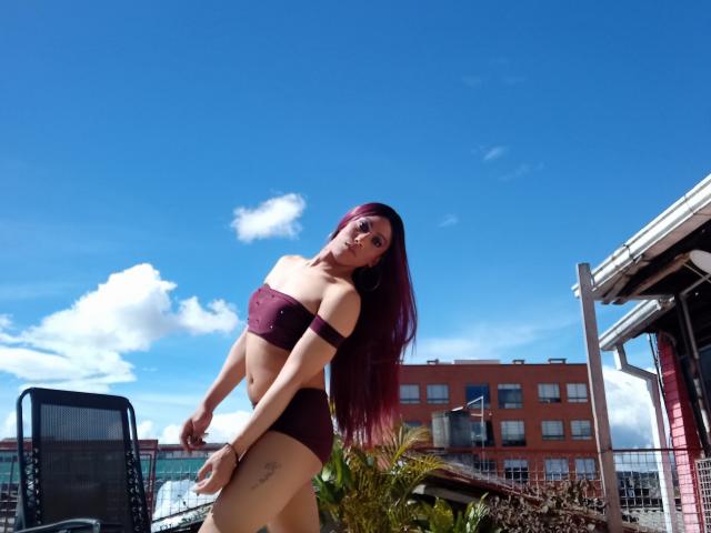 Violetta Gill mit langen, roten Haaren, die einen burgunderfarbenen Bikini trägt und ihre Brüste und ihren Hintern zeigt, steht draußen, blickt nach unten, der Himmel ist klar blau, die Sonne scheint, und im Hintergrund ist ein Gebäude zu sehen..
