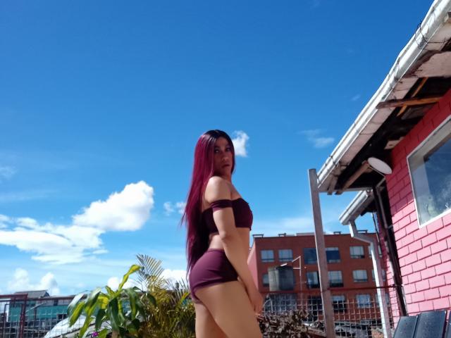 Violetta Gill mit langen, lila Haaren, einem lila Top mit einem Schulterauschnitt und lila Shorts, die ihr Gesäß zeigen, steht draußen vor einem blauen Himmel, einem roten Gebäude und einer grünen Pflanze im Hintergrund..