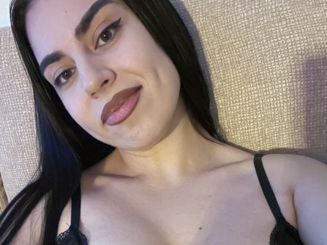 Josephina: Mit langen, schwarzen Haaren, dunklen Augenbrauen, dicker Eyeliner und pinkem Lippenstift, lächelnd mit leicht herausgesteckter Zunge, trägt sie einen schwarzen Spitzen-BH und liegt auf einem strukturierten Sofa..