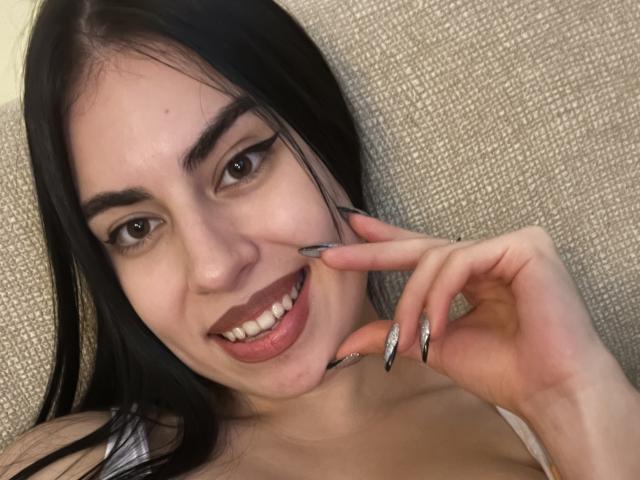JosephinaHot with black hair, brown eyes, and dark lipstick, smiling with a hand touching her cheek, showing off long, shiny, black and silver nails, and her boobs.