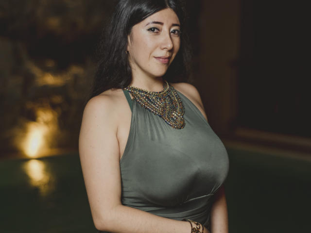 HotPamelaSex with large tits, wearing a green halter top, ornate necklace, and a bracelet, smiling slightly, dark hair, and brown eyes, in a dimly lit, warm-toned setting.