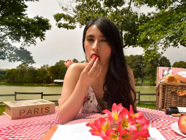 JainaRogers with long black hair, eating a red fruit, wearing a floral dress, sitting at a red-and-white checkered picnic table with pink flowers and a wicker basket.