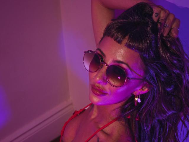 ElizaWells with dark, wavy hair, wearing red lingerie and aviator sunglasses, holding her hair up, glowing with purple and pink lighting, showing a tattoo on her chest.