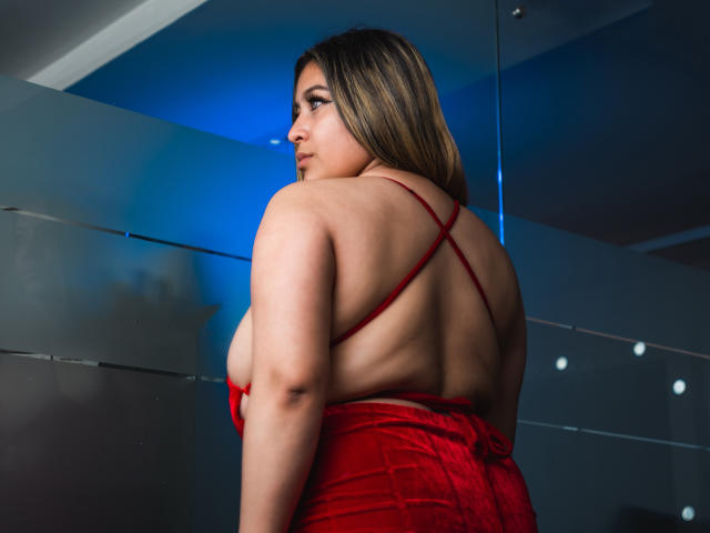 MillyHerder steht mit dem Rücken zur Kamera, trägt ein rotes Spitzenkleid, das ihre großen Brüste und ihren wohlgeformten Hintern zeigt, und blickt über ihre Schulter, wobei im Hintergrund blaues Licht zu sehen ist..