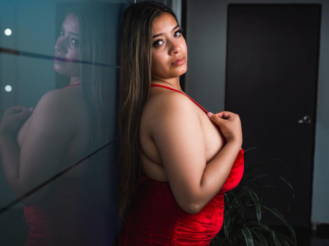 MillyHerder, con cabello largo y castaño, vestida con un vestido rojo, de pie frente a un espejo, sosteniendo sus grandes senos redondos con su mano derecha, con su reflejo visible, mirando a la cámara..