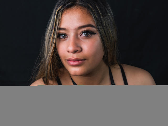 Os olhos de MillyHerder e parte do seu rosto são visíveis acima de uma barreira cinza, horizontal. Ela tem cabelos longos com mechas loiras e está olhando diretamente para a câmera..