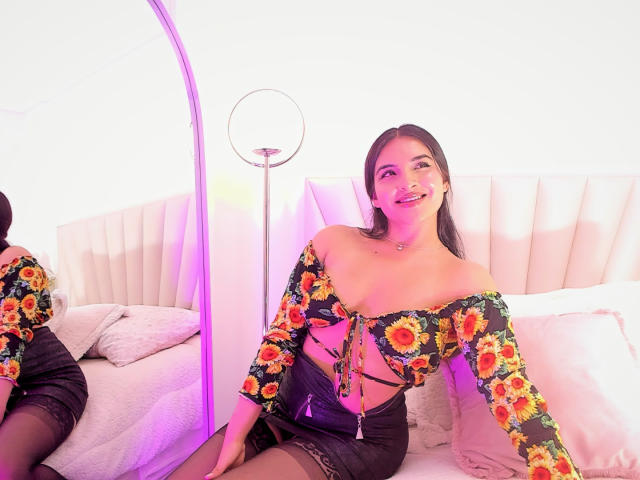 EmilyJensenn with sunflower-patterned top, showing her tits, black skirt, and black stockings, sitting on bed, smiling, in front of mirror, with purple light and ring lamp.