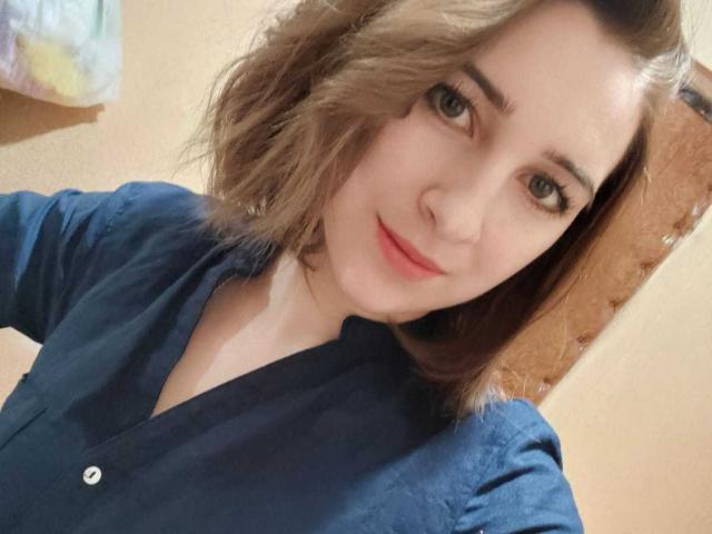 FionaJazz mit welligem, braunem Haar, blauen Augen und einem leichten Lächeln, trägt ein dunkelblaues Hemd und zeigt ein süßes Gesicht sowie einen kleinen Hinweis auf ihre Brüste..