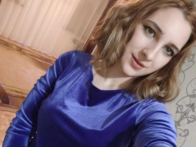 FionaJazz, mit mittelgroßer Brust, trägt ein glänzendes, blaues Samintop. Sie hat hellbraunes, welliges Haar, grüne Augen und ein leichtes Lächeln. Der Hintergrund ist unscharf..